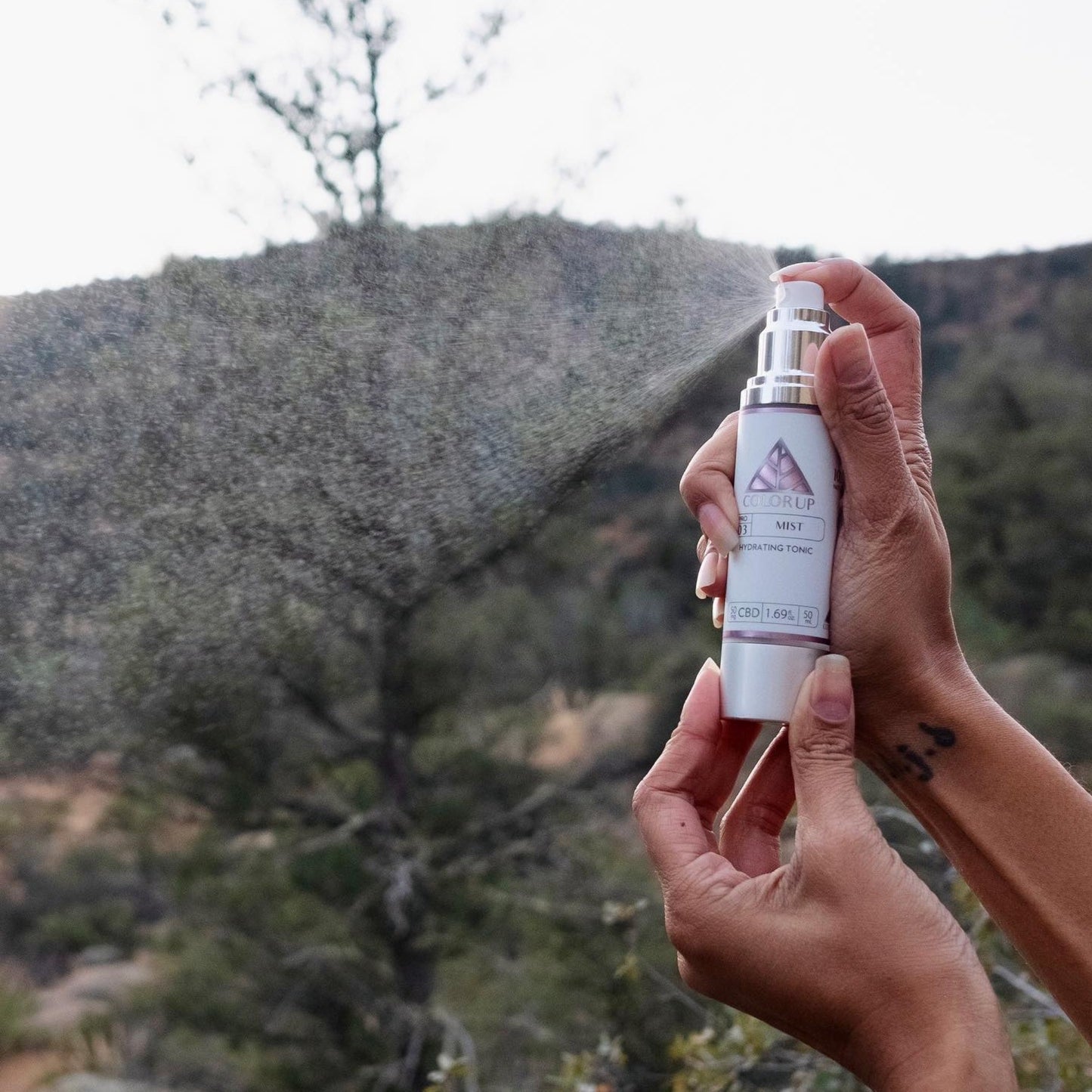 Hands holding Color Up Mist Hydrating Tonic spraying in the forefront with a mountainous background.