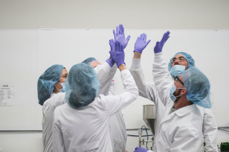 Several Color Up lab scientists in white lab coats, blue hairnets, and blue gloves high-five in a clean indoor setting with a white wall and some equipment visible in the background.