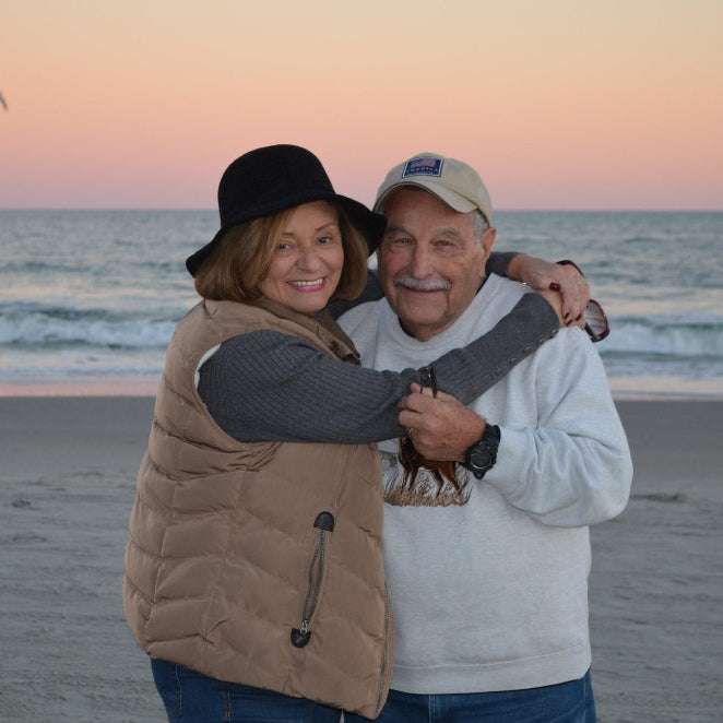 Ellen and Phil Blanch, Color Up advisors, embracing on a beach with a sunset and ocean in the background.